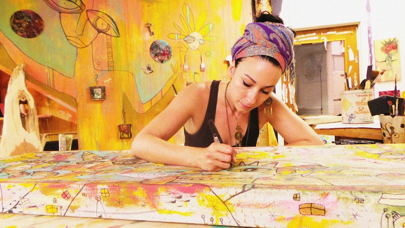 Puede incluir: Una mujer con un pa&ntilde;uelo de cabeza colorido est&aacute; trabajando en un lienzo grande y colorido. El lienzo est&aacute; cubierto de dise&ntilde;os y patrones abstractos. La mujer est&aacute; usando un marcador negro para dibujar en el lienzo.