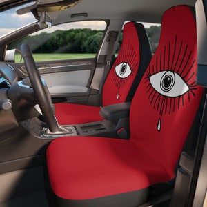 May include: Two red car seat covers with a black outline of a large eye with eyelashes and a single teardrop. The background is red.