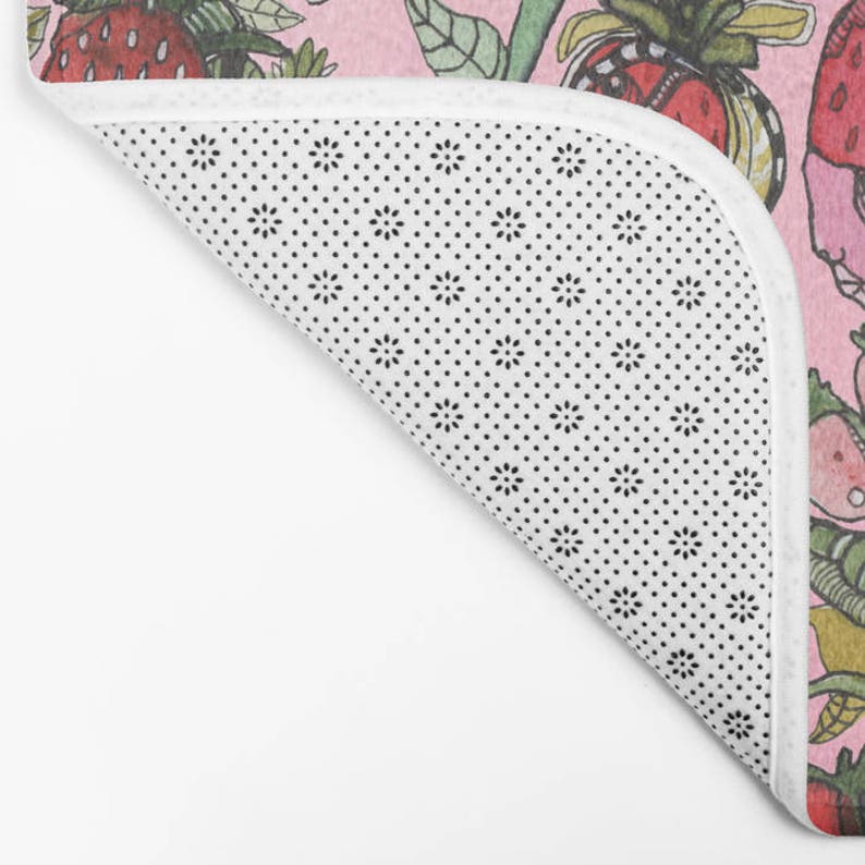 Puede incluir: Una alfombra de ba&ntilde;o rosa con un patr&oacute;n repetido de fresas, hojas e insectos. La alfombra tiene un respaldo antideslizante blanco.