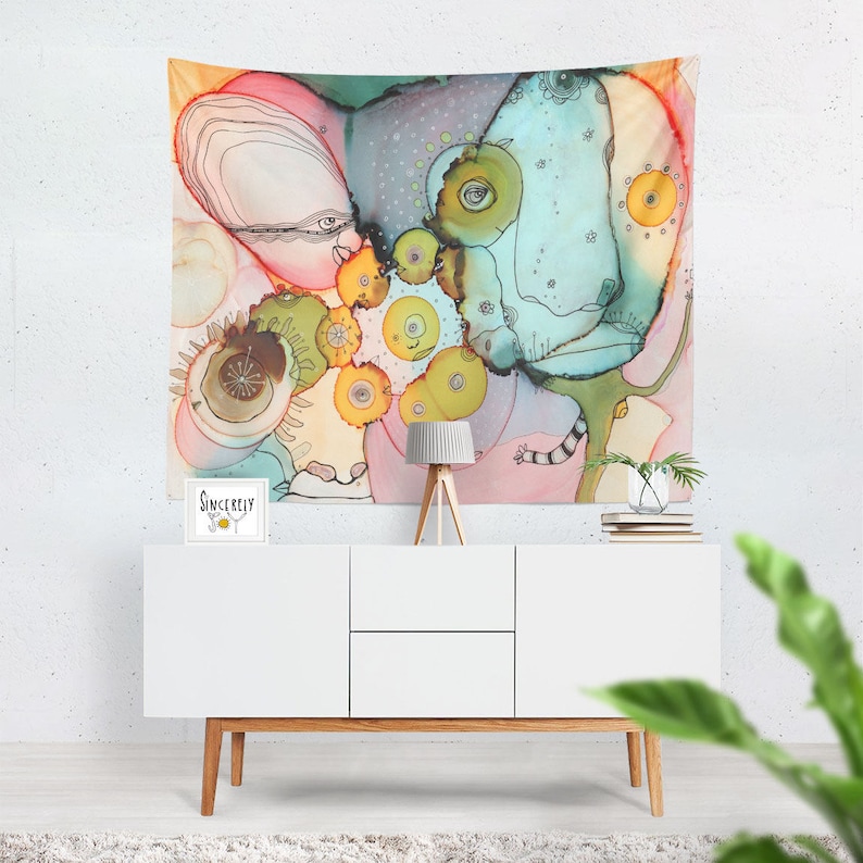 May include: Abstract art tapestry with a colorful design. The artwork features a mix of shapes and colors, including pink, blue, yellow, and green. The tapestry is hanging above a white cabinet with wooden legs. A small lamp and a framed print that says "Sincerely Joy" are on the cabinet.