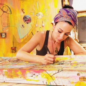 Puede incluir: Una mujer con un pa&ntilde;uelo de colores est&aacute; trabajando en una gran pintura colorida. La pintura est&aacute; sobre una superficie de madera y tiene una variedad de formas y colores. La mujer est&aacute; usando un marcador negro para agregar detalles a la pintura.