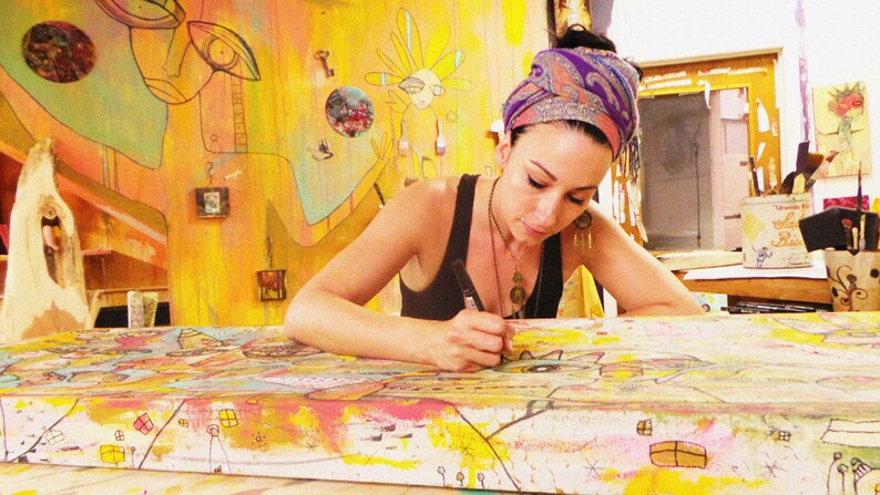 Puede incluir: Una mujer con un pa&ntilde;uelo de colores est&aacute; trabajando en una gran pintura colorida. La pintura est&aacute; sobre una superficie de madera y tiene una variedad de formas y colores. La mujer est&aacute; usando un marcador negro para agregar detalles a la pintura.