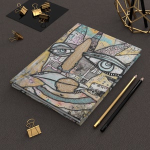 May include: A notebook with a colorful abstract design featuring a face with two eyes and a mouth. The design is made up of geometric shapes, lines, and patterns. The notebook is closed and lying on a gray surface.