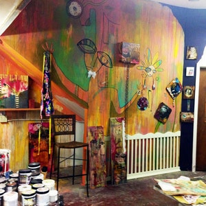May include: A colourful, abstract mural with a large, green, human-like figure painted on the wall. The mural is in a room with a wooden floor and a white railing. There are several small paintings and other artwork on the walls. A wooden chair sits in front of the mural.
