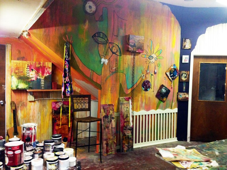 May include: A colourful, abstract mural with a large, green, human-like figure painted on the wall. The mural is in a room with a wooden floor and a white railing. There are several small paintings and other artwork on the walls. A wooden chair sits in front of the mural.