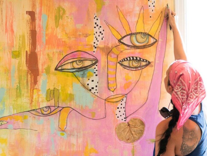 Puede incluir: Obra de arte abstracta que presenta un rostro estilizado con m&uacute;ltiples ojos, en tonos rosa, amarillo y naranja. La obra incluye detalles de l&iacute;neas negras y un elemento de hoja. Se ve a una persona con un pa&ntilde;uelo rosa trabajando en el arte.