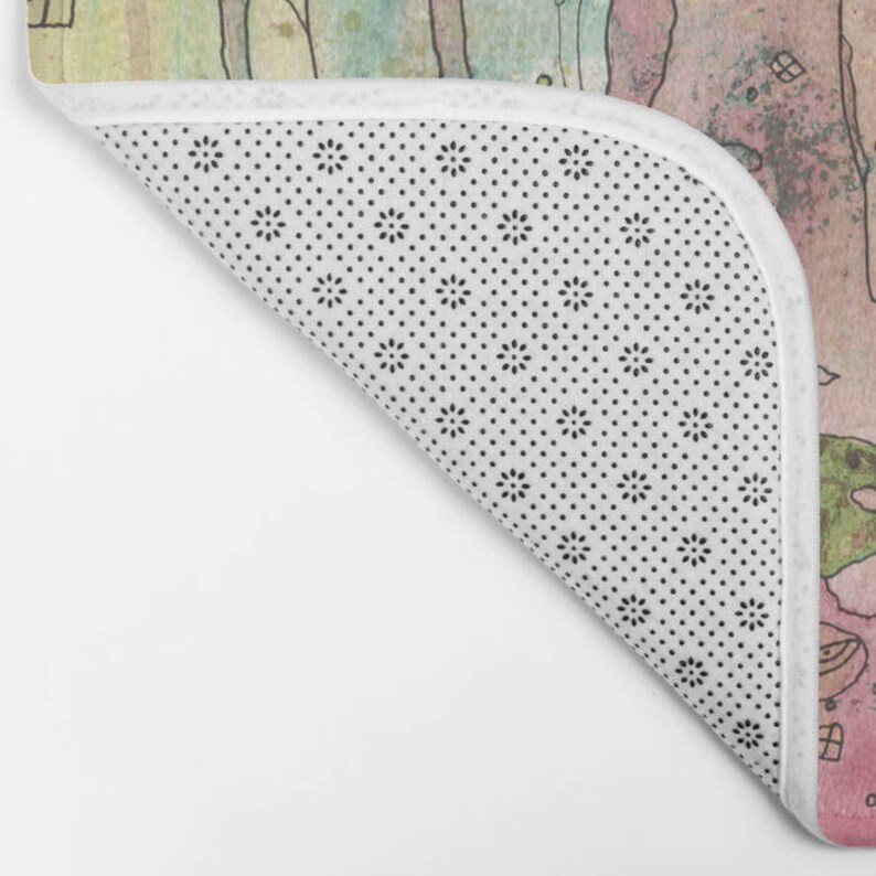 Peut inclure: Un tapis de bain en aquarelle avec un design fantaisiste repr&eacute;sentant un paysage rose et vert avec des arbres et des maisons. Le tapis a un dos antid&eacute;rapant blanc avec un motif de petits points noirs.