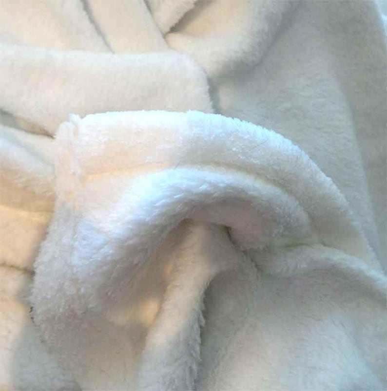 May include: Close-up of a white plush fabric with a soft, fuzzy texture. The fabric is folded and draped, showing the texture and softness.