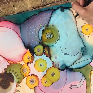 May include: Abstract artwork featuring a whimsical creature with a light blue head, yellow eyes, and a green body. The artwork uses a combination of pink, yellow, and teal colors. A hand with a pen is visible, adding a sense of creation.