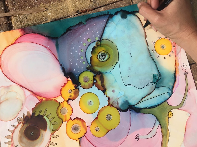 May include: Abstract artwork featuring a whimsical creature with a light blue head, yellow eyes, and a green body. The artwork uses a combination of pink, yellow, and teal colors. A hand with a pen is visible, adding a sense of creation.