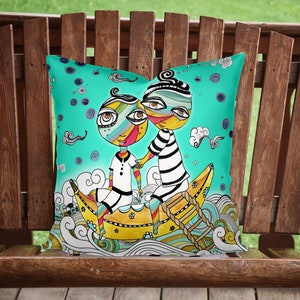 May include: A turquoise pillow with a whimsical illustration of two cartoon characters riding a yellow banana-shaped boat on a turquoise sea. The characters have large eyes and colorful features. The background is filled with clouds and swirls.
