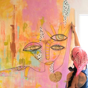 May include: A large abstract painting with a pink background and a colourful face with multiple eyes. The painting is being hung on a wall by a person wearing a pink bandana.