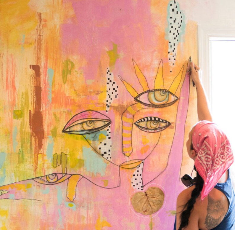 May include: A large abstract painting with a pink background and a colourful face with multiple eyes. The painting is being hung on a wall by a person wearing a pink bandana.