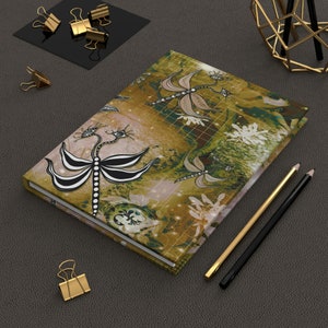 May include: A green and gold journal with a dragonfly design. The dragonfly is black and white with a stylized design. The journal has a textured cover and a white spine.