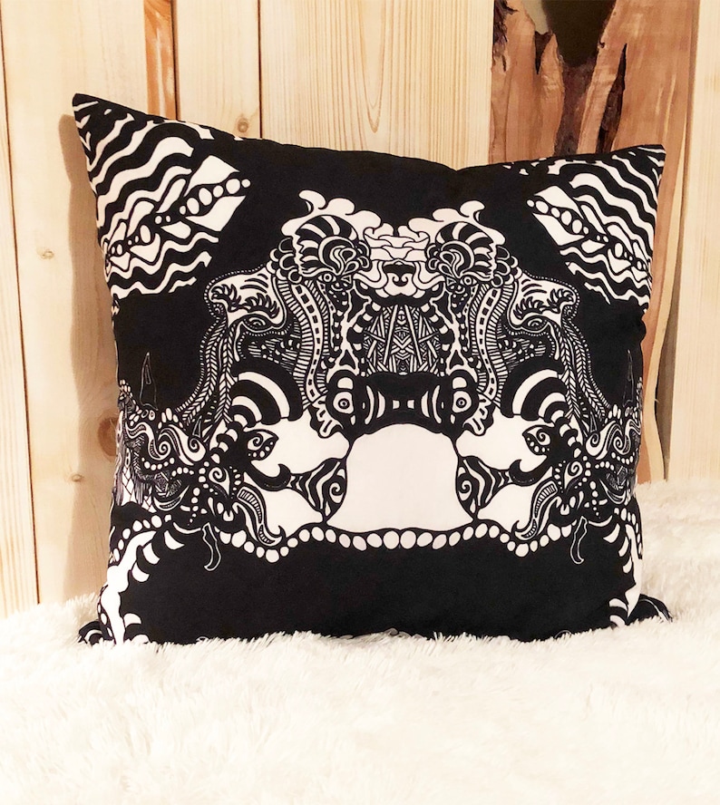 May include: Black and white decorative pillow with an intricate design featuring a stylized face. The pillow is made of fabric and has a square shape.
