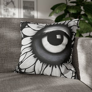 May include: Square decorative pillow featuring a black and white eye design. The eye is the focal point, surrounded by a floral pattern. The pillow is placed on a gray sofa, creating a striking contrast.