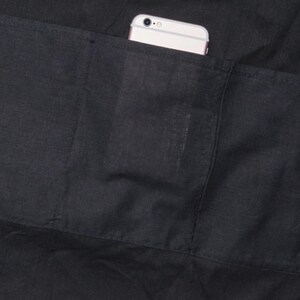 Puede incluir: Primer plano de un delantal negro con un bolsillo grande. Un tel&eacute;fono inteligente blanco es parcialmente visible en el bolsillo. El delantal est&aacute; hecho de una tela oscura con costuras visibles.