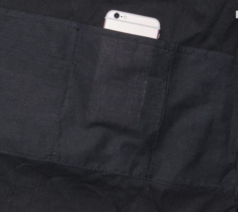 Puede incluir: Primer plano de un delantal negro con un bolsillo grande. Un tel&eacute;fono inteligente blanco es parcialmente visible en el bolsillo. El delantal est&aacute; hecho de una tela oscura con costuras visibles.