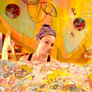 May include: A woman with a colorful headscarf is working on a large canvas painting. The canvas is covered in colorful abstract faces and patterns. The woman is smiling and appears to be enjoying her work.