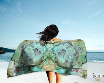 May include: A woman wearing a large, colourful scarf with a whimsical, abstract design. The scarf features a repeating pattern of eyes and circles in shades of green, blue, and brown. The woman is standing on a beach with the sea in the background.