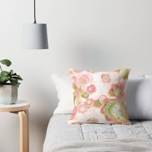 May include: A white throw pillow with a pink and green abstract pattern. The pillow is on a white bed with a gray blanket.
