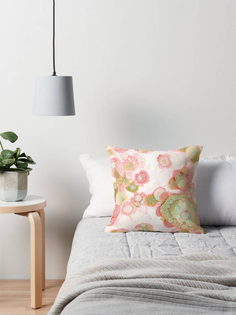 May include: A white throw pillow with a pink and green abstract pattern. The pillow is on a white bed with a gray blanket.