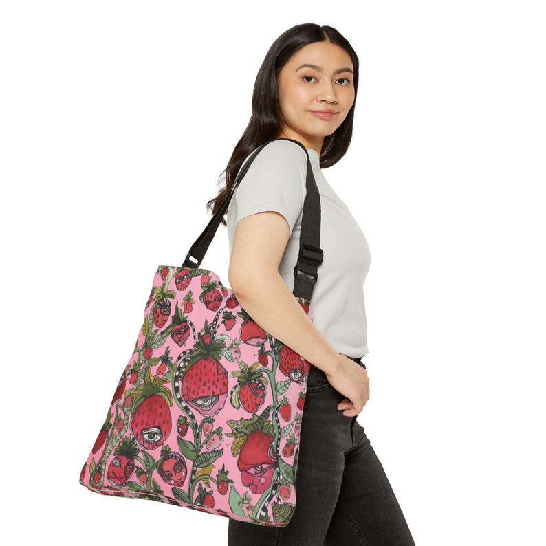 Puede incluir: Un bolso tote rosa con un patr&oacute;n repetido de fresas estilizadas con caras y ojos. Las fresas son rojas con hojas y tallos verdes. El bolso tiene una correa negra para el hombro.