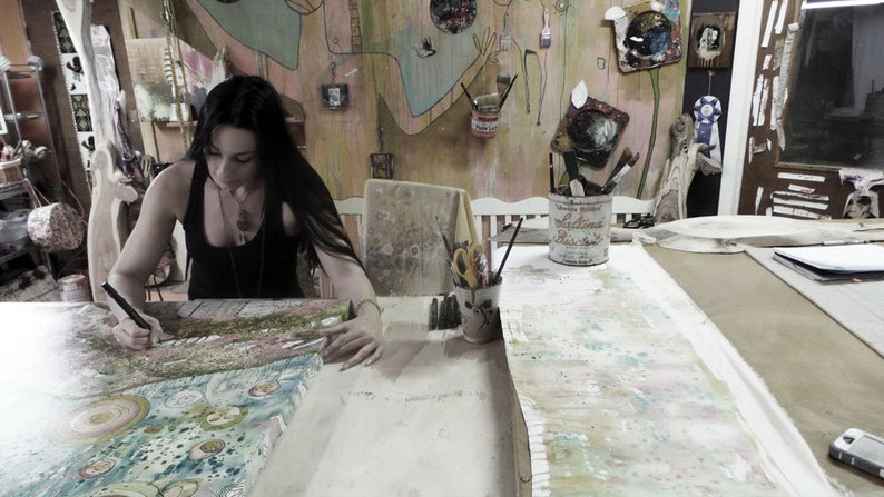 May include: A woman with long dark hair is working on a large canvas painting in her art studio. The canvas is covered in colorful paint and patterns. There are various art supplies and tools on the table, including paintbrushes, scissors, and a tin can with the text "Hollandaise Blue Satin" on it.