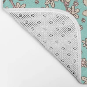 May include: A turquoise blue bath mat with a white background and a pattern of pink and white flowers. The mat has a white non-slip backing with small black dots.