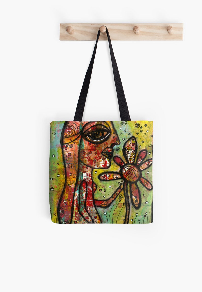 Peut inclure: Un sac fourre-tout color&eacute; avec une peinture d'une personne avec un grand &oelig;il et une fleur. La personne est peinte dans des tons de rouge, orange et jaune, tandis que la fleur est rouge et jaune. L'arri&egrave;re-plan est un m&eacute;lange de vert, jaune et blanc.