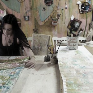 May include: A woman with long dark hair is working on a large canvas painting in her art studio. The canvas is covered in colorful paint and patterns. There are various art supplies and tools on the table, including paintbrushes, scissors, and a tin can with the text "Hollandaise Blue Satin" on it.