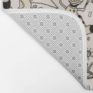 May include: A beige bath mat with a black and white pattern of stylised figures. The mat has a non-slip backing with a small black dot pattern.