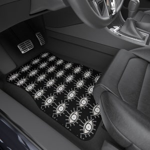 May include: Black car floor mat with a white eye pattern. The mat is in the driver's side footwell of a car.