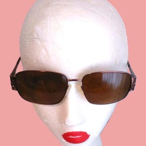 May include: Brown sunglasses with a rectangular frame and dark lenses. The frame is a metallic brown color with decorative detailing on the temples. The sunglasses are displayed on a white mannequin head with red lipstick.