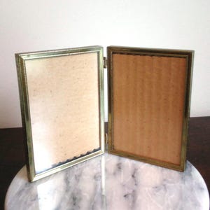 May include: A double-hinged picture frame with a metallic gold finish and a beaded edge detail. The frame consists of two rectangular panels connected by a central hinge, each featuring a clear glass front and a light brown cardboard backing.