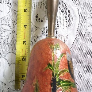 Pu&ograve; includere: Campana vintage in ottone con finitura smaltata rosso-arancio, con un motivo floreale con accenti verdi e neri. La campana misura circa 7.6 cm di altezza, con manico e bordo in ottone. Un metro a nastro &egrave; accanto alla campana.