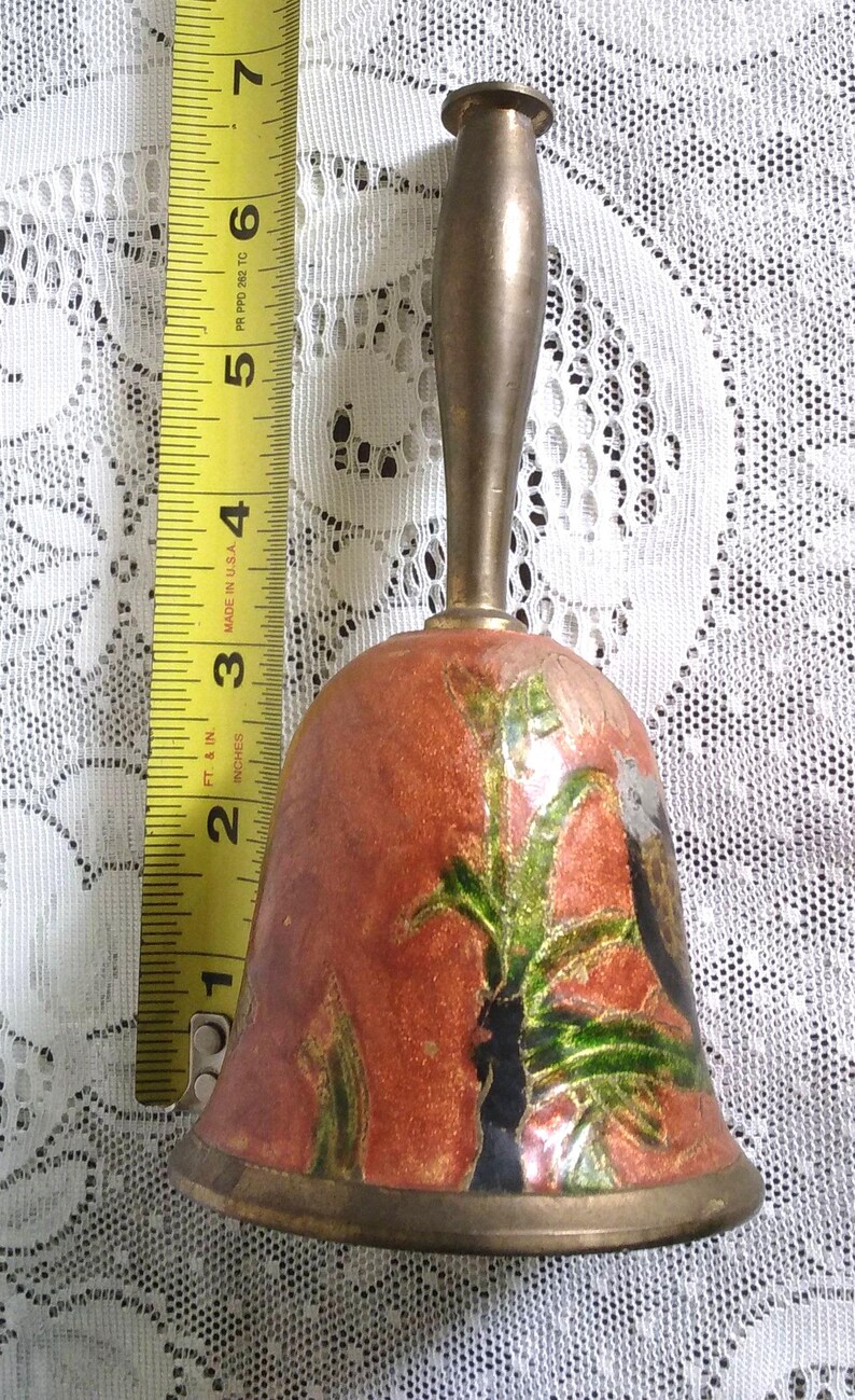 Pu&ograve; includere: Campana vintage in ottone con finitura smaltata rosso-arancio, con un motivo floreale con accenti verdi e neri. La campana misura circa 7.6 cm di altezza, con manico e bordo in ottone. Un metro a nastro &egrave; accanto alla campana.