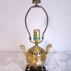 May include: A brass table lamp base shaped like a samovar with two side handles, a central light socket, and a harp attachment topped with a small brass bird figurine. The lamp rests on a black, carved wooden base with four small feet.
