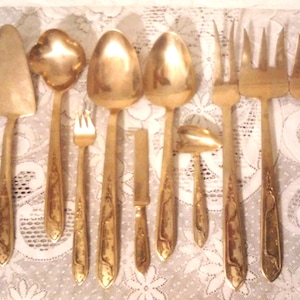 May include: A collection of gold-colored flatware, including knives, cake servers, spoons, forks, and a ladle, arranged on a white lace tablecloth. The utensils have decorative detailing on the handles, suggesting a vintage or antique style.