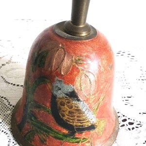 Pu&ograve; includere: Campana in ottone vintage con finitura smaltata rossa. La campana presenta una dettagliata illustrazione di un uccello appollaiato tra le foglie. Il manico e la base della campana sono in ottone.