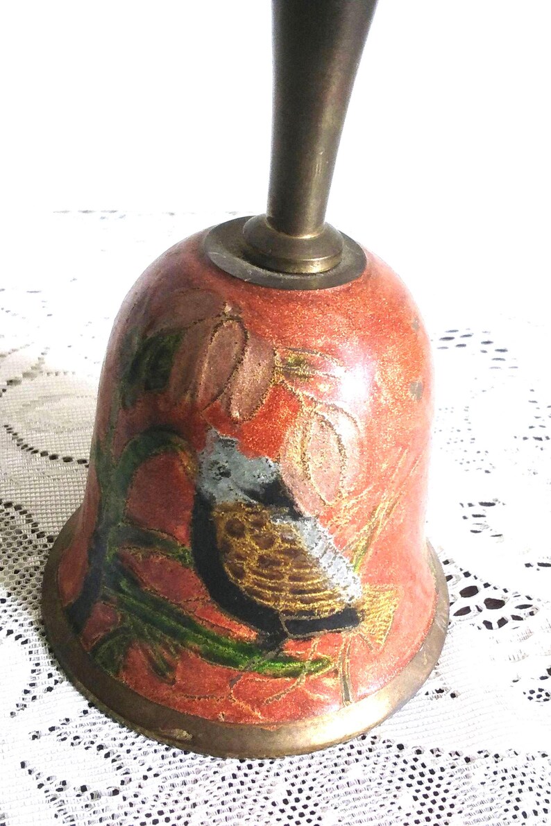 Pu&ograve; includere: Campana in ottone vintage con finitura smaltata rossa. La campana presenta una dettagliata illustrazione di un uccello appollaiato tra le foglie. Il manico e la base della campana sono in ottone.