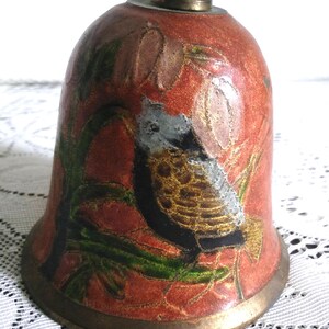 Pu&ograve; includere: Campana vintage con finitura smaltata rossa e un design dettagliato di uccelli e fiori. La campana presenta un manico e una base in ottone, con accenti verdi, dorati e neri. La campana ha una forma arrotondata, con una base svasata.