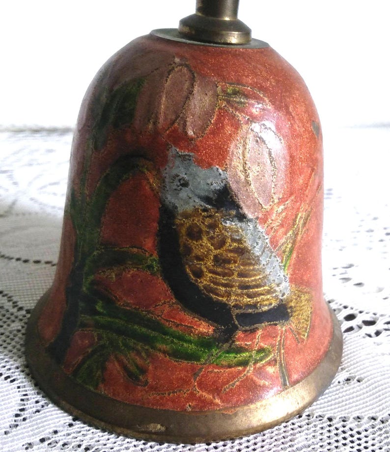Pu&ograve; includere: Campana vintage con finitura smaltata rossa e un design dettagliato di uccelli e fiori. La campana presenta un manico e una base in ottone, con accenti verdi, dorati e neri. La campana ha una forma arrotondata, con una base svasata.