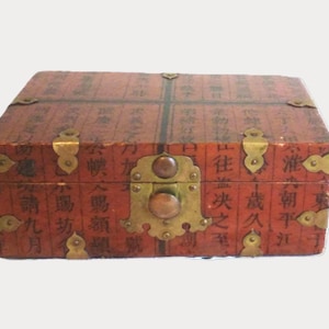 May include: A rectangular wooden box with a reddish-brown finish, featuring black Chinese calligraphy characters across the surface. The box is accented with decorative brass corner plates and a central brass latch mechanism with a circular knob.
