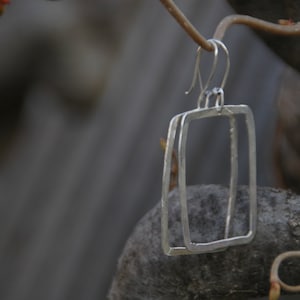 May include: Silver earrings with a rectangular shape. The earrings are made of two thin pieces of metal that are layered on top of each other.