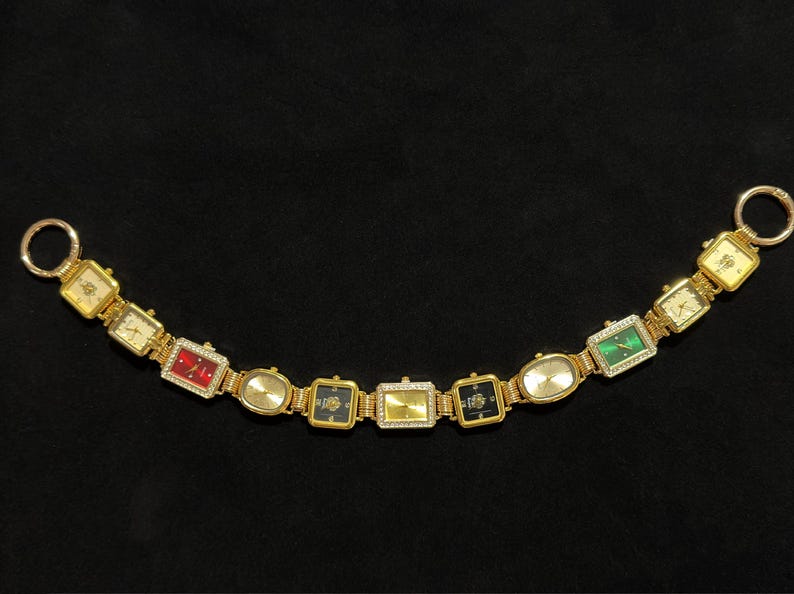 May include: A gold-toned watch bracelet with multiple rectangular and oval watch faces. The watch faces feature various colors, including red, green, and black, and are adorned with small crystals. The bracelet has two circular clasps.