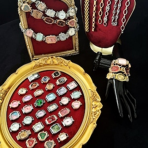 Può includere: Una collezione di orologi quadrati colorati con cinturini di diversi colori, esposti su uno sfondo di velluto rosso in una cornice dorata. Gli orologi sono anche esposti su un vassoio dorato con un interno in velluto rosso. Ci sono anche diverse catene dorate e argentate esposte su un supporto in velluto rosso.