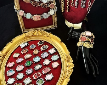 Könnte beinhalten: Eine Sammlung von bunten quadratischen Uhren mit verschiedenen farbigen Armbändern, die auf einem roten Samt-Hintergrund in einem goldenen Rahmen ausgestellt sind. Die Uhren werden auch auf einem goldenen Tablett mit einem roten Samt-Innenraum ausgestellt. Es gibt auch mehrere goldene und silberne Ketten, die auf einem roten Samt-Ständer ausgestellt sind.