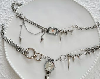 May include: Two silver wristwatches with chain link bands and various silver charms, including safety pins, spikes, and a padlock. The watches are displayed on a white plate.