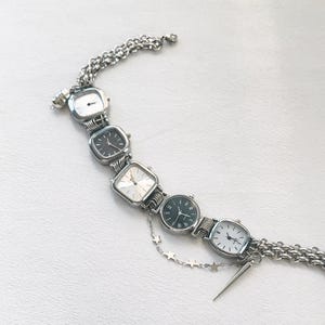 May include: A silver bracelet with five square-shaped watches attached. The watches have silver bands and black or white faces. A silver chain with star charms hangs from the bracelet.
